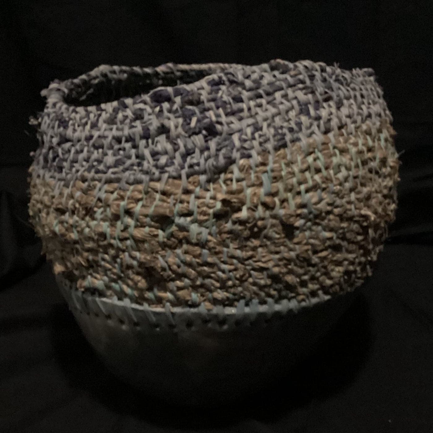 Tall coiled fibre vessel over a burnished clay base, photographed against black