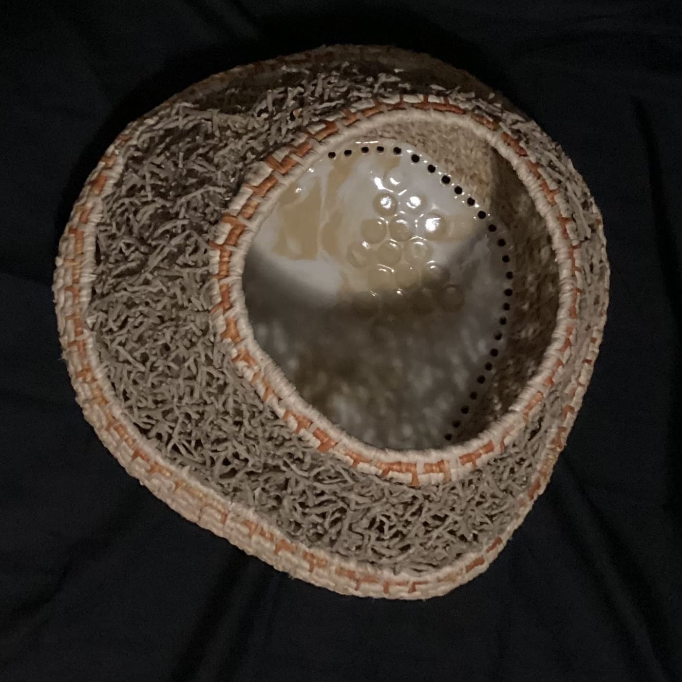 Coiled fibre vessel with a glazed disc set into its base, photographed against black